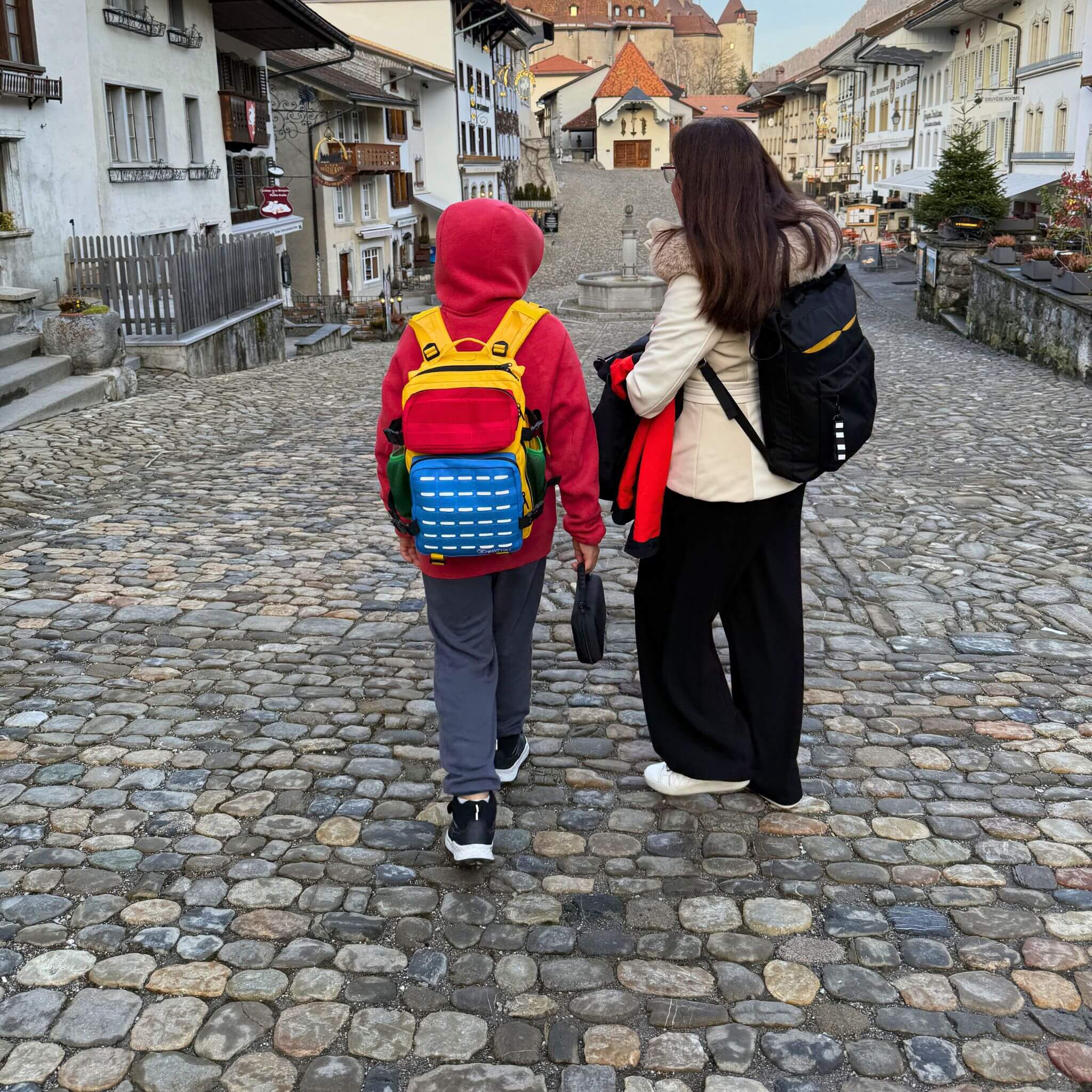 Boy wearing colourful backpack exploring Swiss village with his mum