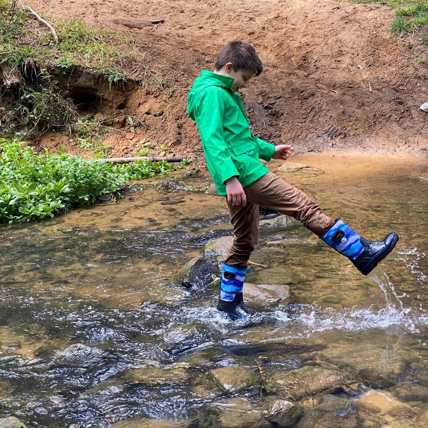 Boy playing in stream wearing rainwear by Wavetree