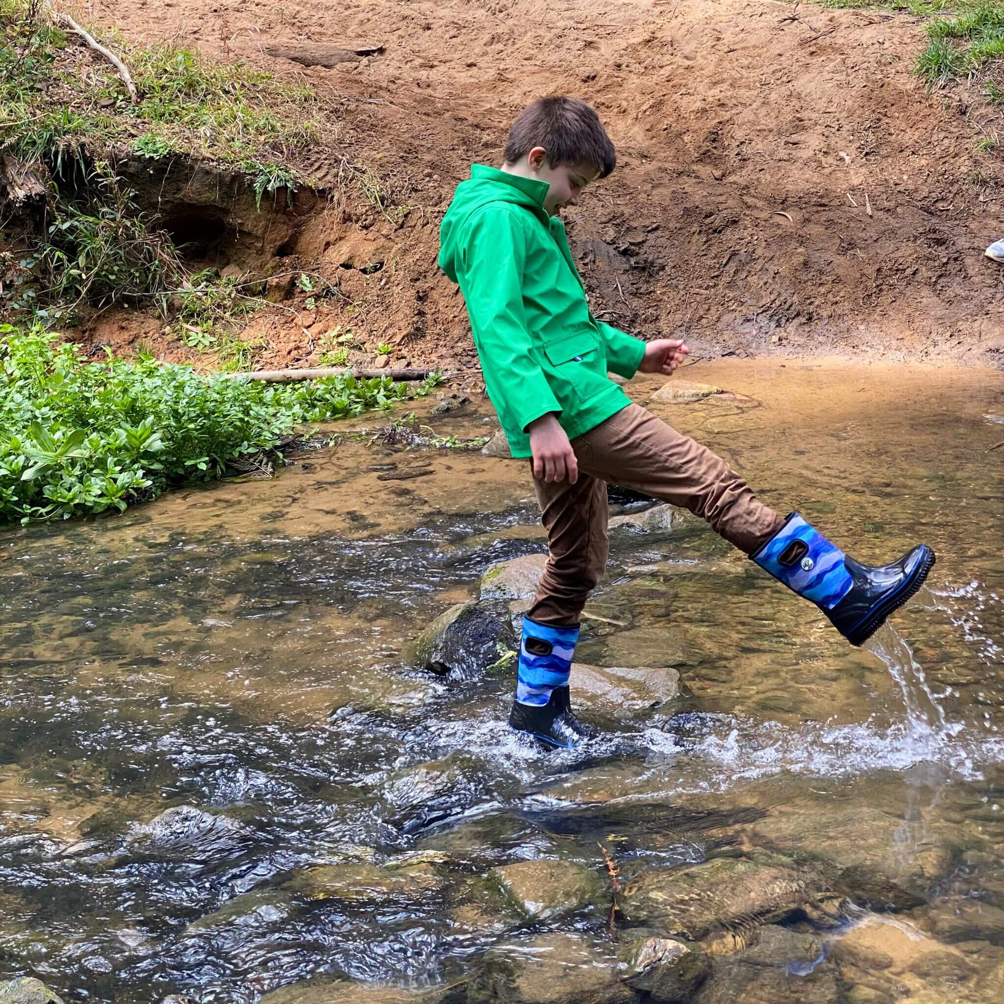 Boy playing in stream wearing rainwear by Wavetree