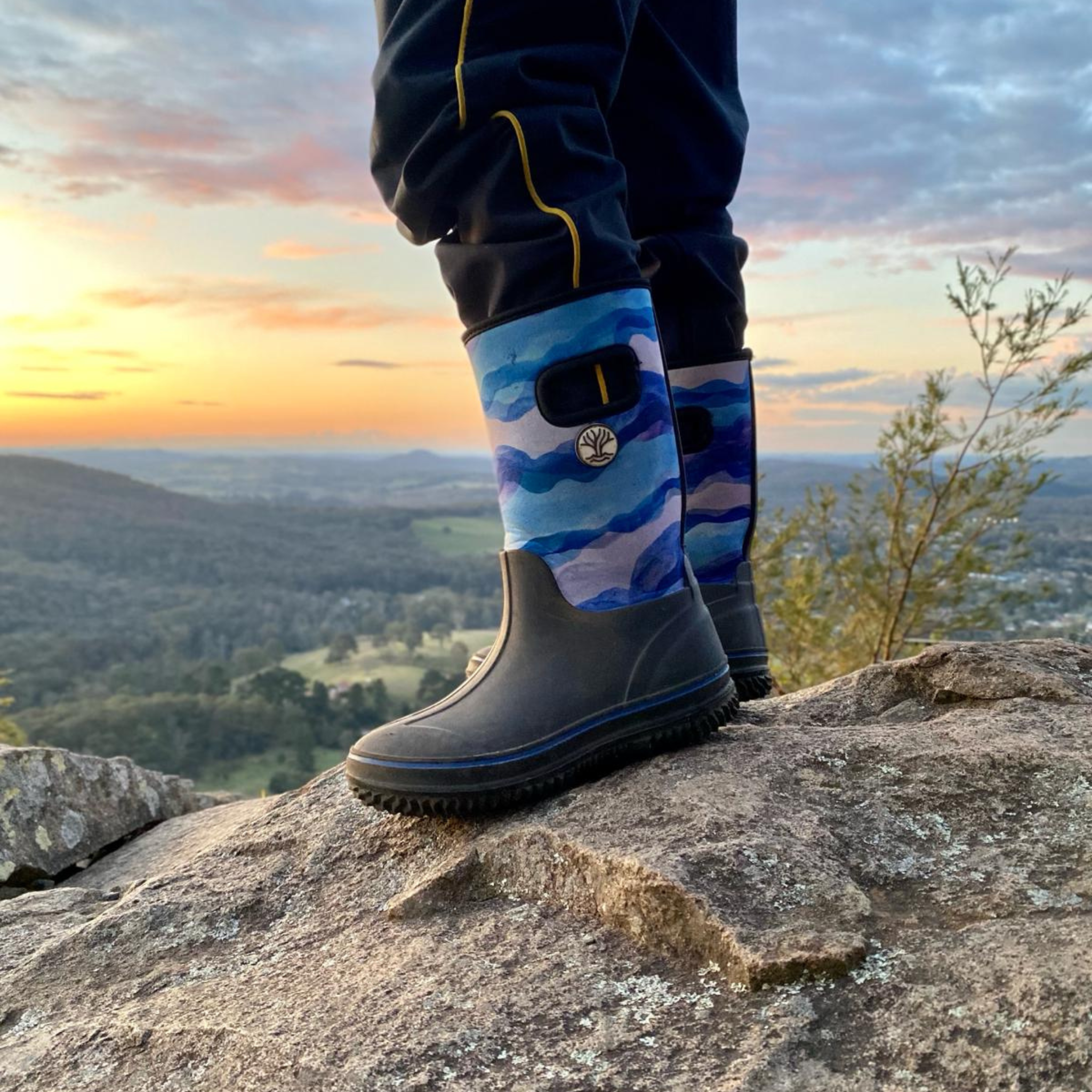 Child in blue rain boots gumboots stood on rock with sunset view in background