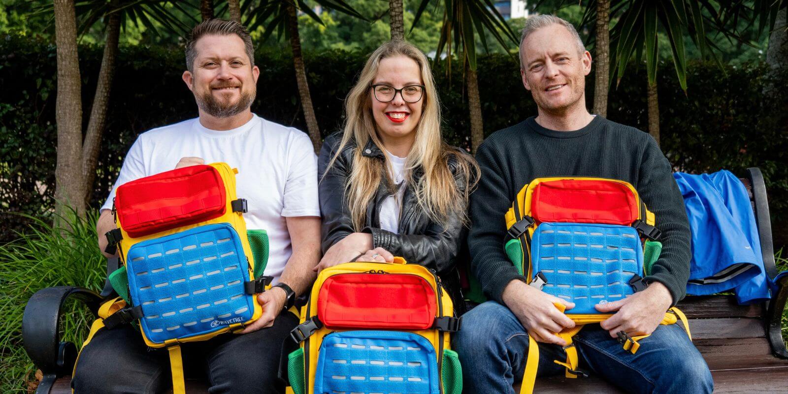 Three people sitting on a bench with colorful backpacks in a garden setting